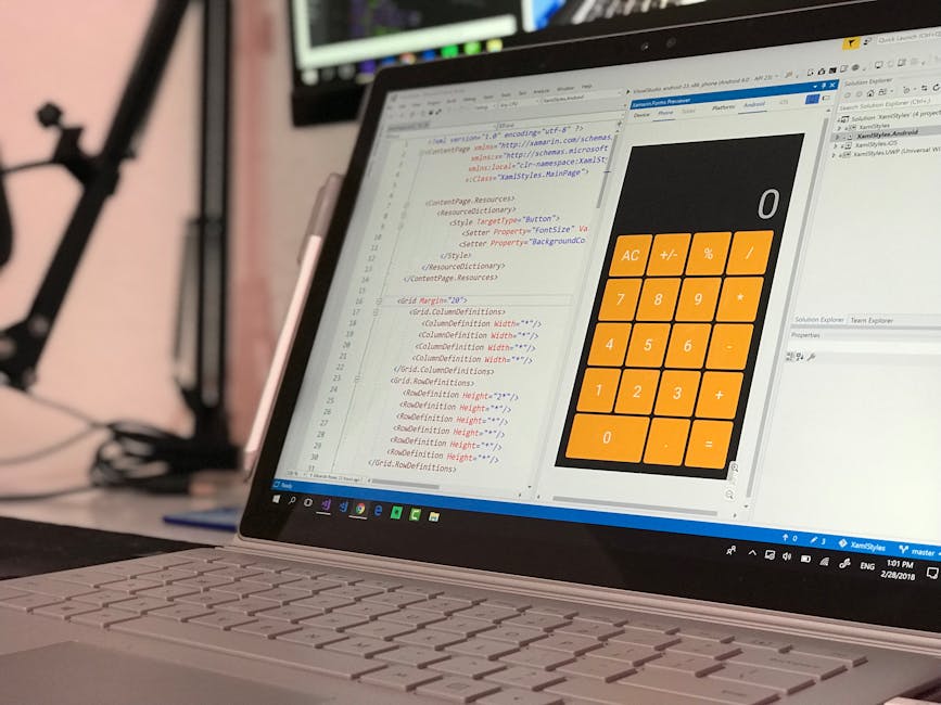A close-up image of a laptop computer displaying a software development environment and a large calculator application with orange buttons on the screen. The laptop is placed on a desk with a black ke