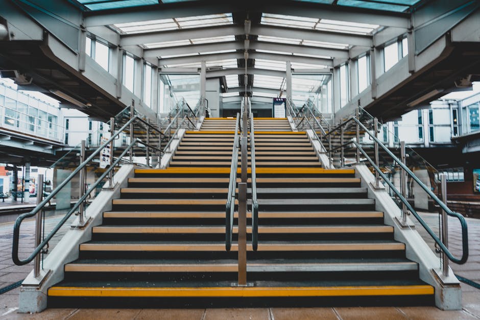 A downward view of a staircase inside a tunnel or underground passage, with steps made of wood with anti-slip strips on each treads. The staircase is flanked by metal handrails on both sides, attached